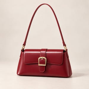 Könnte beinhalten: Eine tiefrote Schultertasche mit glatter, strukturierter Oberfläche. Die Tasche verfügt über einen einzelnen Tragegriff und eine Frontklappe, die mit einer goldfarbenen Metallschnalle verschlossen wird. Das Design ist rechteckig mit abgerundeten Ecken.