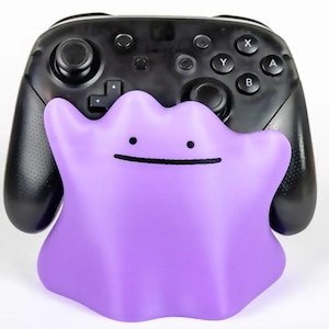 Könnte beinhalten: Ein schwarzer kabelloser Videospiel-Controller hinter einem violetten, amorphen, klecksförmigen Ständer. Der Ständer hat ein einfaches Gesicht mit zwei kleinen schwarzen Punkten als Augen und einem horizontalen schwarzen Strich als Mund.