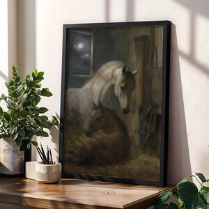 May include: A framed wall art print depicting a white horse standing over a brown foal resting in a dimly lit stable. A small window in the background shows a night sky with a full moon. The artwork is enclosed in a simple black rectangular frame.