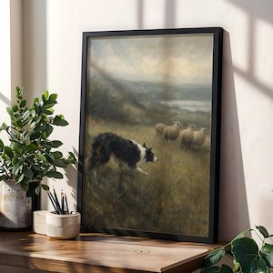 May include: A framed wall art print depicting a black and white border collie herding a small group of sheep in a grassy, overcast field. The painting features muted earth tones, rolling hills in the distance, and a soft, cloudy sky above the scene.