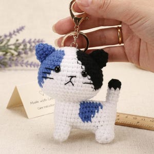 Op de afbeelding: Een gehaakte katten-sleutelhanger met een wit lichaam, een blauw linkeroor, een zwart rechteroor en een blauwe vlek op de zijkant. De kat heeft zwarte geborduurde ogen en snorharen, een kleine zwarte mond en een staart met een zwart uiteinde.