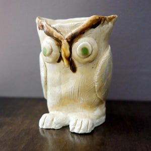 May include: A cream-colored ceramic owl figurine with a textured surface, featuring large protruding eyes with green centers, a brown-painted brow line, and carved wing details. The owl has a hollow top opening and stands on two feet.
