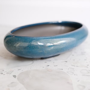 May include: An oval-shaped ceramic planter with a glossy, deep blue glaze on the exterior and a smooth, dark gray interior. The vessel features a low-profile design with rounded edges and a wide, open top, showcasing a reflective finish.
