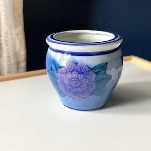 May include: A small, round ceramic planter featuring a white interior and a blue exterior. The exterior is decorated with a central purple and pink floral motif surrounded by green leaves, accented by a dark blue rim at the top and base.