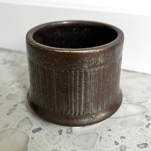 May include: A small, cylindrical ceramic vessel with a dark, metallic brown glaze. The exterior features a vertical ribbed texture around the body, with a smooth, slightly flared base and a plain, circular rim at the top opening.