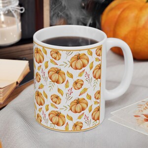 May include: A white ceramic mug featuring a repeating pattern of orange pumpkins, yellow autumn leaves, and small red berries. The mug is filled with a dark liquid and is framed by a thin gold border, showcasing a seasonal harvest theme.