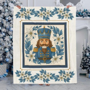 May include: A rectangular throw blanket featuring a central illustration of a nutcracker in a blue uniform, surrounded by a wreath of white poinsettias and blue holly leaves. The design is framed by a gold border and floral accents.