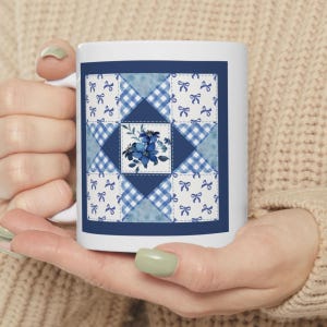 May include: A white ceramic mug featuring a square quilt-inspired graphic. The design includes alternating patches of blue and white gingham, solid blue triangles, and white squares with blue bows, centered with a blue floral illustration.