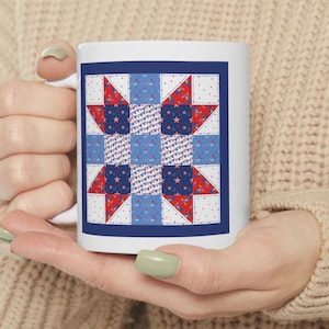 May include: A white ceramic mug featuring a square graphic print of a patriotic quilt pattern. The design consists of a grid of red, white, and blue squares with star motifs, bunting, and polka dots, all framed by a solid dark blue border.