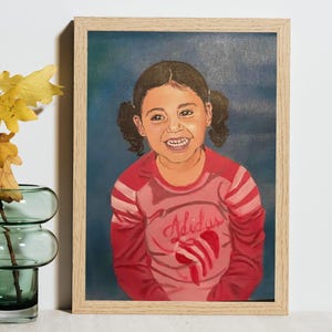 May include: A framed oil painting on canvas depicting a smiling child with dark hair in pigtails. The child wears a pink long-sleeved shirt with the word Adidas and a red heart graphic printed on the front. The painting is set in a light wood frame.