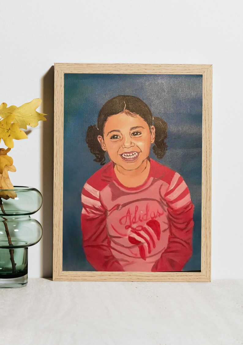 May include: A framed oil painting on canvas depicting a smiling child with dark hair in pigtails. The child wears a pink long-sleeved shirt with the word Adidas and a red heart graphic printed on the front. The painting is set in a light wood frame.