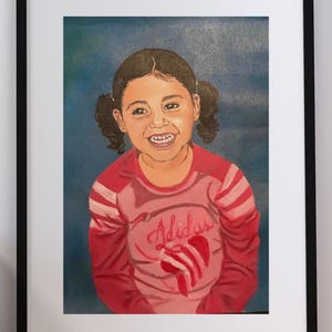May include: A framed painting of a smiling child with dark hair in pigtails, wearing a long-sleeved pink and red shirt with the word Adidas and a red graphic logo printed on the front, set against a solid dark blue background.