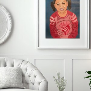 May include: A framed portrait painting of a smiling child with dark hair in pigtails. The child wears a long-sleeved pink shirt with red stripes on the sleeves and a red heart graphic on the chest. The background is a solid dark blue color.