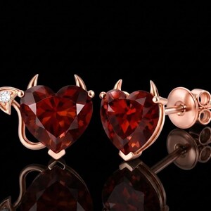 Puede incluir: Un par de pendientes de botón en oro rosa con granates rojos en forma de corazón. Cada pendiente presenta pequeños cuernos de diablo en la parte superior y una cola curva en el lateral, con una de las colas decorada con una pequeña piedra clara.