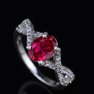 May include: A silver ring featuring a central oval-cut deep red gemstone held by four prongs. The band displays a crisscross design on both sides, encrusted with small, clear, sparkling stones that extend along the shoulders of the ring.