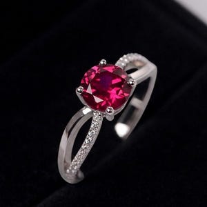 Op de afbeelding: Een zilverkleurige ring met een ronde, gefacetteerde, dieprode middensteen die door vier pootjes wordt vastgehouden. De band heeft een gespleten ontwerp met aan één kant een rij kleine, heldere, fonkelende steentjes en een gladde afwerking.