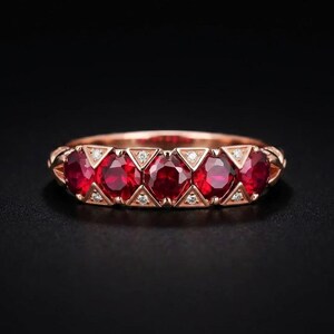 May include: A rose gold ring featuring five round-cut deep red rubies arranged in a row. Between each ruby, small triangular settings contain tiny white diamonds. The band has a polished finish and a consistent width across the front.