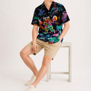 Puede incluir: Una camisa de manga corta con botones sobre un fondo negro que presenta un vibrante estampado gráfico de neón de un gato con gafas de sol, sosteniendo bebidas y surfeando, rodeado de palmeras, medusas y elementos del espacio exterior.