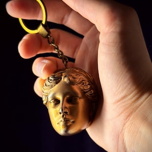 Op de afbeelding: Een goudkleurige metalen sleutelhanger met een gebeeldhouwd menselijk hoofd met gedetailleerde gelaatstrekken en golvend haar. De hanger is bevestigd aan een ronde splitring door een korte, gedraaide metalen ketting, wat een samenhangend metalen accessoire vormt.