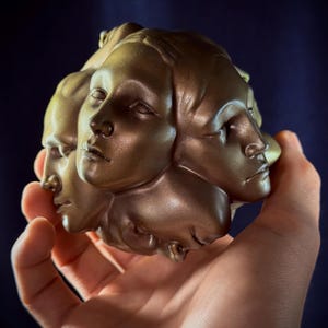 Op de afbeelding: Een bolvormig beeldhouwwerk met meerdere menselijke gezichten in het oppervlak verwerkt. Het object heeft een metallic bronskleurige afwerking met gladde contouren en gedefinieerde gelaatstrekken zoals ogen, neuzen en lippen in een complexe vorm.