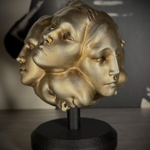 Pode incluir: Uma escultura de cor dourada apresentando múltiplos rostos humanos dispostos em um grupo esférico. Os rostos têm olhos fechados e expressões neutras. A escultura está montada em um poste cilíndrico preto e uma base circular preta.