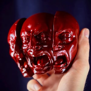 Pode incluir: Uma escultura vermelho-escura apresentando três rostos humanos idênticos dispostos em fila, cada um com a boca bem aberta e os olhos fechados. Os rostos são segmentados em três partes distintas, criando um efeito em camadas com acabamento liso.