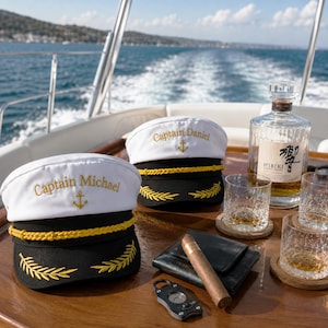 Może przedstawiać: Dwie biało-czarne czapki kapitańskie ze złotym haftem z imionami Captain Michael i Captain Daniel, szklana butelka whisky Hibiki, dwie kryształowe szklanki z bursztynowym płynem, czarny skórzany portfel i obcinacz do cygar.