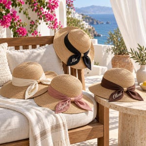 May include: Four straw sun hats with wide brims and ribbon bands tied in bows. The ribbons are black, cream, dusty rose, and dark brown. Each ribbon features a name embroidered in white script: Mia, Emma, Sophie, and Olivia, respectively.