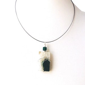 May include: A wire-wrapped pendant necklace featuring a rectangular, off-white stone with a dark green stone accent. The pendant is suspended from a thin, black wire choker. A small, green cube-shaped bead sits above the rectangular stone.