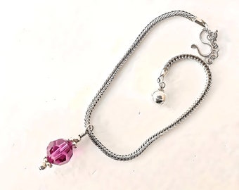 May include: A silver chain anklet with a purple crystal bead and a silver charm. The anklet has a lobster clasp and a small silver bell.