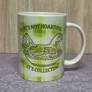 May include: White ceramic mug with a green plaid pattern and a brown illustration of a hen in a nest. The mug has the text "IT'S NOT HOARDING" above the hen and "IT'S COLLECTING" below.