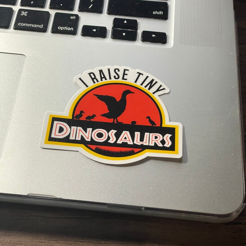 May include: A white sticker with a red and yellow dinosaur silhouette. The sticker says "I raise tiny dinosaurs" with a goose and goslings inside the dinosaur silhouette.