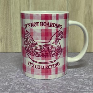 May include: White ceramic mug with a pink gingham pattern and a pink illustration of a hen in a nest. The mug has the text "IT'S NOT HOARDING" above the hen and "IT'S COLLECTING" below.
