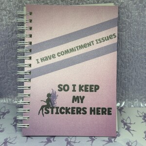May include: A purple and gray spiral bound notebook with the text "I have commitment issues so I keep my stickers here" and a silhouette of a fairy with wings.