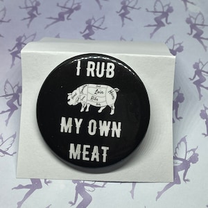 May include: A black round button with white text that reads "I RUB MY OWN MEAT." A diagram of a pig with labeled cuts of meat is in the center. The button is on a white surface.