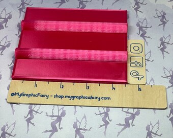 May include: A bright pink plastic 3D printed mold with three slots for making resin crafts. The mold is approximately 15 inches long and is sitting on a white surface with a pattern of fairies.