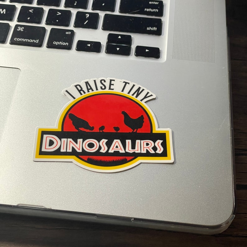 May include: A sticker with a red, yellow, and black Jurassic Park-style logo that says "I raise tiny dinosaurs". The logo features a silhouette of three chickens, two chicks, and a green patch of grass.