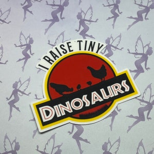 May include: A white sticker with a red, yellow, and green Jurassic Park-style logo. The logo says "I raise tiny dinosaurs" with a silhouette of a mother chicken and chicks inside.