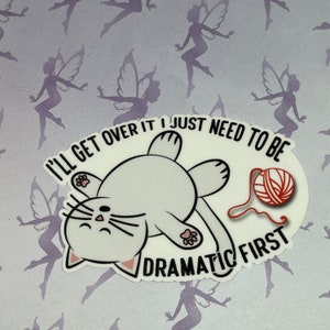 May include: White cat sticker with a red ball of yarn. The cat is lying on its back with its paws in the air. The text reads "I'll get over it I just need to be dramatic first."