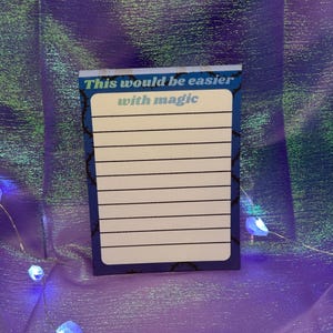 May include: A rectangular notepad with a dark blue border featuring a black thorn pattern. The top section displays the text "This would be easier" in white and "with magic" in light blue. The center contains white lined paper for writing.