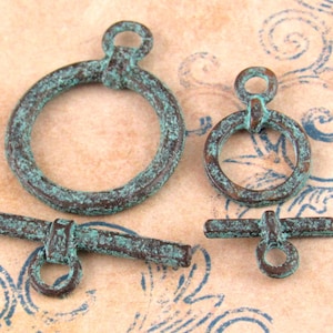 May include: Two toggle clasps for jewelry making, each consisting of a circular ring and a T-shaped bar. The metal components feature a weathered, oxidized finish with a dark brown base and a textured, verdigris green patina throughout.