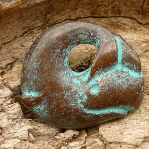May include: A small, bronze-coloured pendant shaped like a stylised animal with a hole in the centre. The pendant has a turquoise patina and is resting on a piece of wood.
