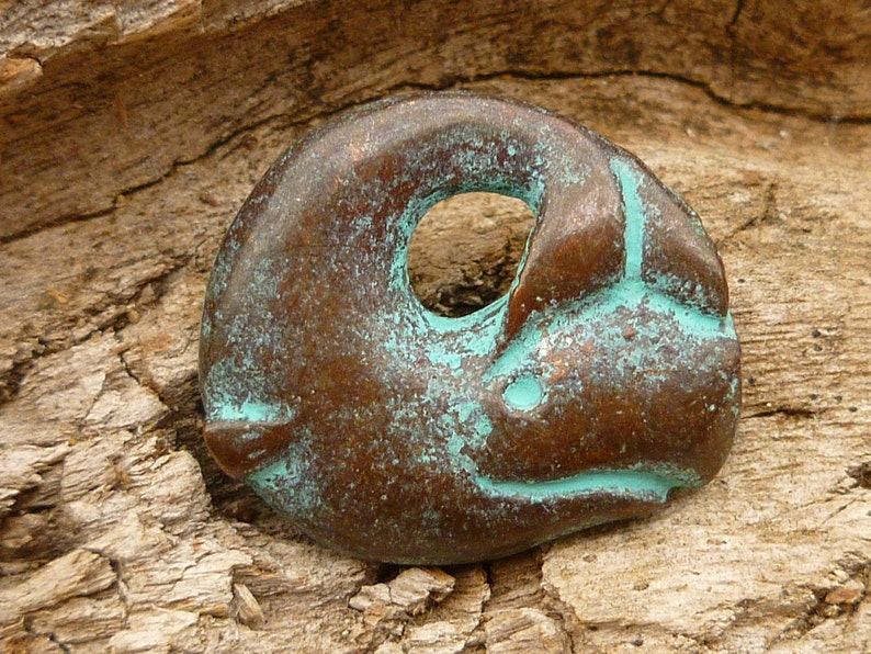 May include: A small, bronze-coloured pendant shaped like a stylised animal with a hole in the centre. The pendant has a turquoise patina and is resting on a piece of wood.