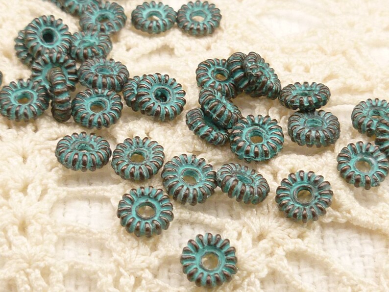 May include: A collection of turquoise-colored, flower-shaped beads with a distressed, antique finish. The beads have a central hole and a detailed, ridged outer edge. They are scattered on a cream-colored, open-weave fabric.