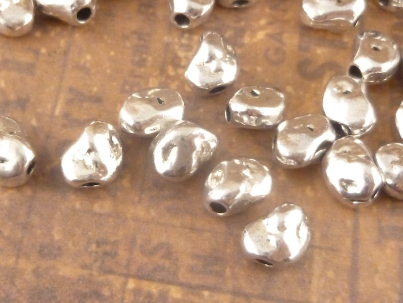 May include: A collection of silver-toned metal beads with a rough, irregular texture. The beads are small and have a hole in the center for stringing.