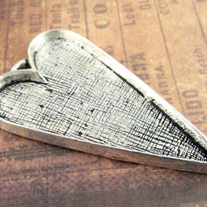 May include: A silver heart-shaped pendant with a textured surface. The pendant is designed to hold a small image or photo.