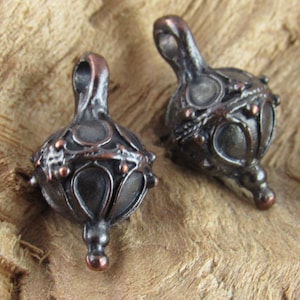 May include: Two ornate, dark metal pendants with a teardrop shape and intricate detailing. The pendants have a loop at the top and a small, rounded base. Copper-colored accents add contrast to the dark metal. The pendants are likely for jewelry making.