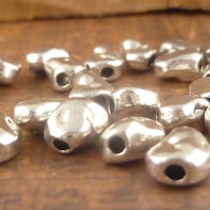 May include: A group of silver heart-shaped beads with a hole in the center. The beads are scattered on a brown surface.