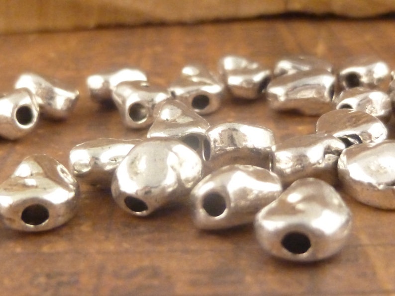 May include: A group of silver heart-shaped beads with a hole in the center. The beads are scattered on a brown surface.