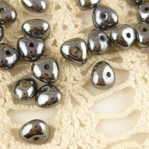 May include: A collection of dark gray, metallic, teardrop-shaped beads with a hole in the center. The beads are scattered on a white crocheted surface.
